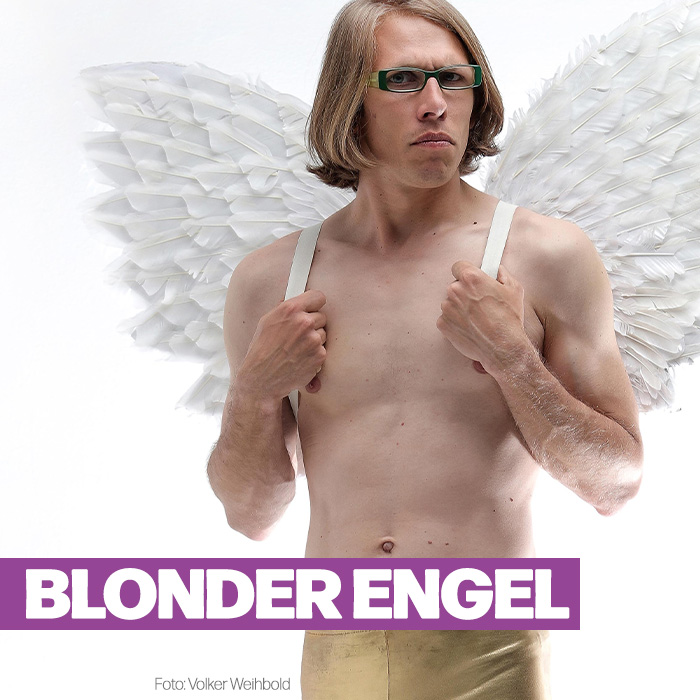 Blonder Engel (Brille und mittellanges Haar) steht vor weißem Hintergrund. Er trägt eine hautenge goldfarbene Hose und nackten Oberkörper. Auf seinem Rücken sind große weiße Flügel befestigt, deren Halterung er vorne wie Hosenträger mit beiden Händen festhält.