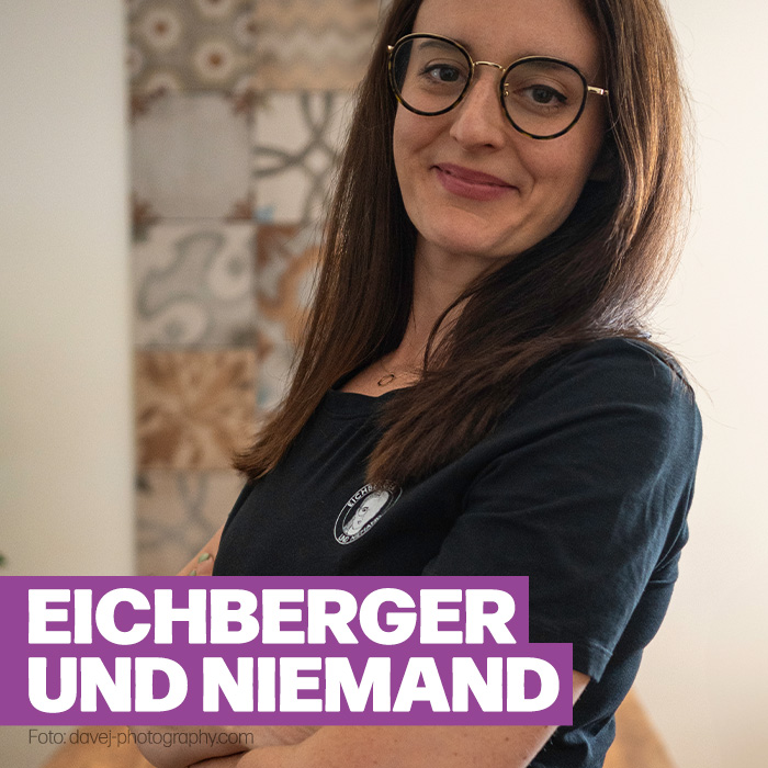 Die Kabarettistin „Eichberger und Niemand“ steht seitlich zur Kamera und schaut in diese mit verschmitztem Lächeln. Sie trägt ein blaues Oberteil, eine Brille und langes dunkles Haar.