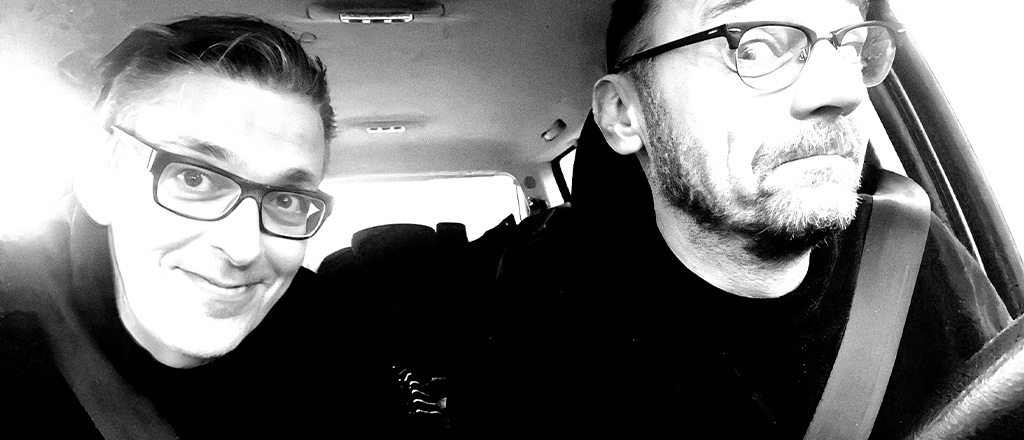 Schwarzweißfoto von Gebrüder Moped im Auto. Martin Moped sitzt am Steuer und blickt nach vorne, Franz Moped sitzt am Beifahrersitz und hat das Selfie aufgenommen. Beide grinsen seitlich in die Kamera.Schwarzweißfoto von Gebrüder Moped im Auto. Martin Moped sitzt am Steuer und blickt nach vorne, Franz Moped sitzt am Beifahrersitz und hat das Selfie aufgenommen. Beide grinsen seitlich in die Kamera.