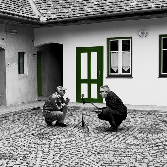 Schwarzweißfoto (mit grünen Farbelementen bei Holztüren im Hintergrund) von Gebrüder Moped in einem Innenhof eines Bauernhauses. Martin Moped links und Franz Moped rechts hocken sich auf einem gepflasterten Boden gegenüber, dazwischen steht ein kleines Stativ mit montiertem Handy.
