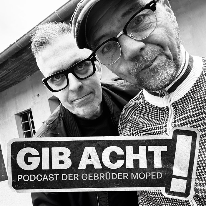 Quadratisches Podcast-Cover: Schwarz-Weiß-Foto der Gebrüder Moped, Aufnahme bis zur Brust. Die beiden stehen nebeneinander und schauen direkt in die Kamera. Auf der linken Seite Franz Moped, schwarz gekleidet, rechts Martin Moped mit Kappe und karierter Weste. Darunter in Form eines abgewetzten Aufklebers der Schriftzug des Podcasts: „Gib Acht! Podcast der Gebrüder Moped“