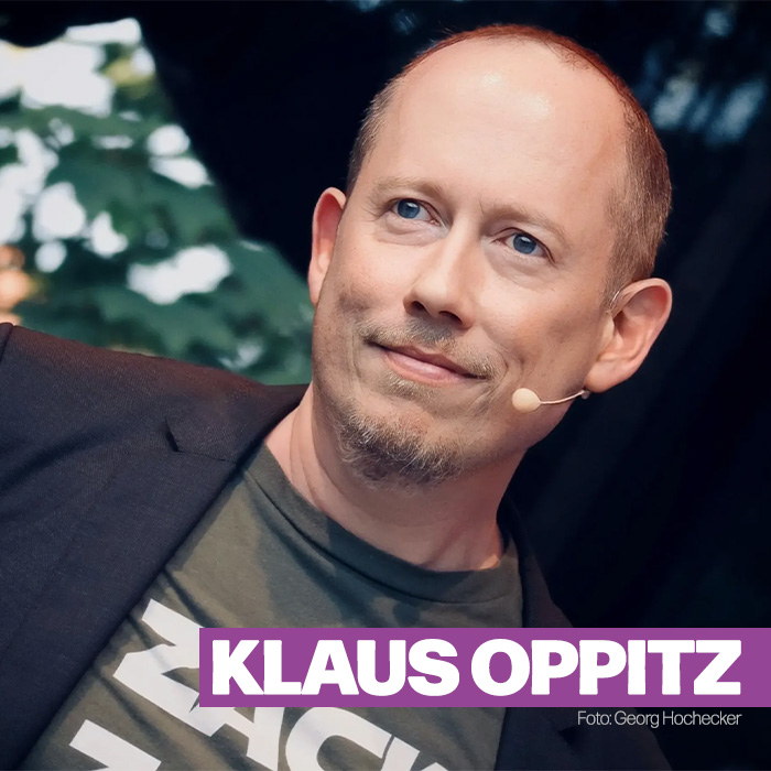 Der Kabarettist Klaus Oppitz ist leicht seitlich geneigt von vorne fotografiert. Das Headset im Gesicht spricht für einen Liveauftritt, im Hintergrund sind Bäume zu sehen. Er trägt Sakko und T-Shirt, hat kurzes Haar und macht einen erfreuten Eindruck.