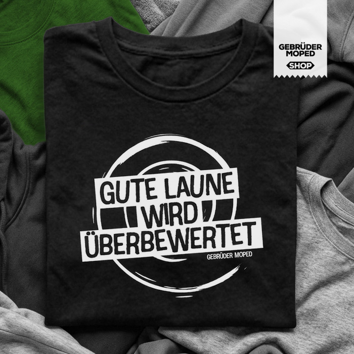 Schwarzweißfoto (mit grünem Farbelement im Hintergrund) eines gefalteten schwarzen T Shirts mit dem Aufdruck „Gute Laune wird überbewertet“ und kleinem „Gebrüder Moped“-Schriftzug darunter. Das Shirt liegt auf mehreren hellen und dunklen Kleidungsstücken, rechts oben hängt ein Etikett mit der Aufschrift „Gebrüder Moped Shop“.