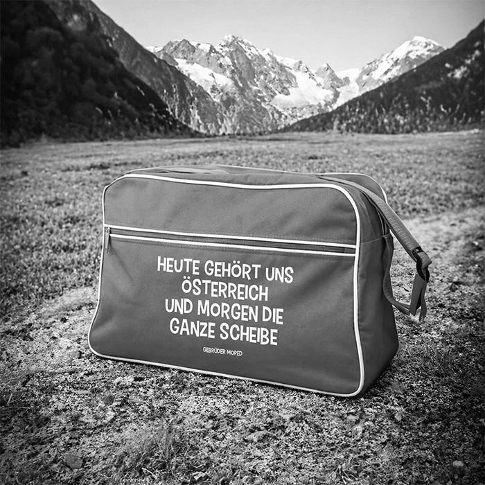 Schwarzweißfoto einer Sporttasche auf einer Wiese vor alpiner Hochgebirgslandschaft. Auf der Tasche steht „Heute gehört uns Österreich und morgen die ganze Scheibe“, klein darunter „Gebrüder Moped“. Im Hintergrund sind Berge mit schneebedeckten Gipfeln zu sehen.